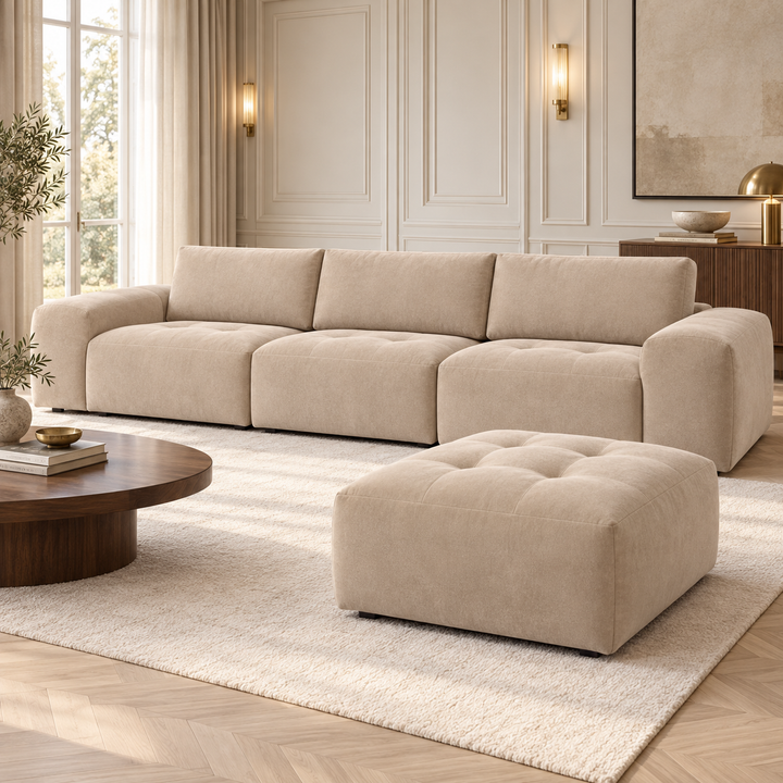 Beige Madele-soffa med tre sittdelar och matchande djuphäftad puff intill ett lågt soffbord i mörkt trä.