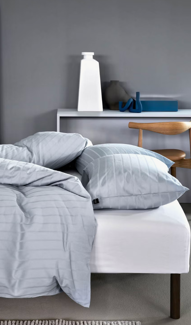 Høie Milano bedding on a bed viewed from the side, with a light blue duvet cover and pillows laid flat; subtle tonal stripes