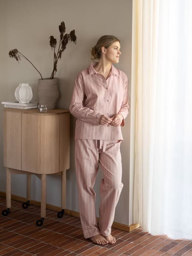 Nainen roosassa raidallisessa Høie Pilen -pyjamassa seisoo ikkunan vieressä