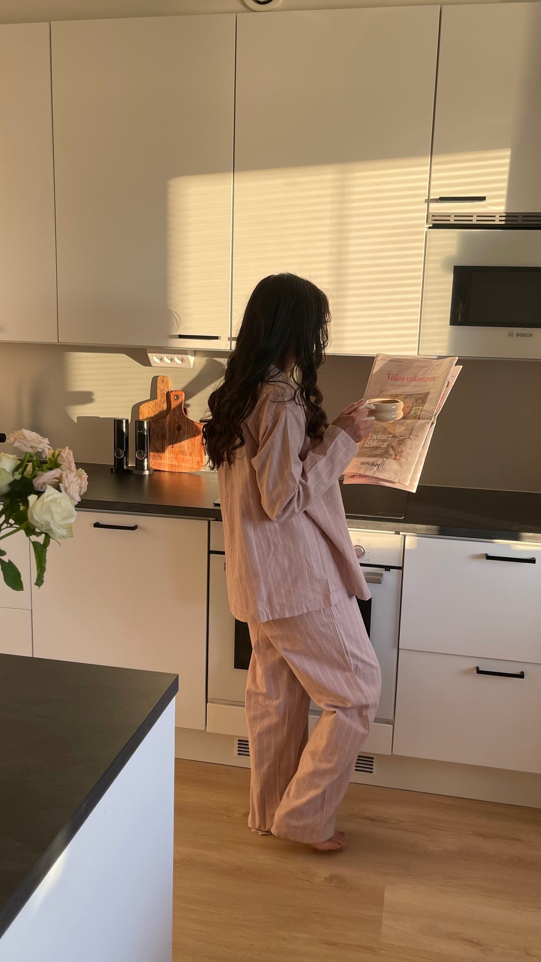 Nainen roosassa Høie Pilen -pyjamassa seisoo keittiössä ilta-auringossa