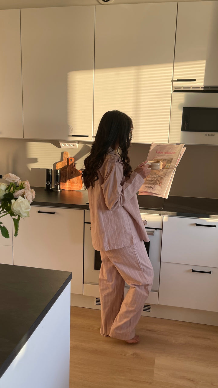Nainen roosassa Høie Pilen -pyjamassa seisoo keittiössä ilta-auringossa