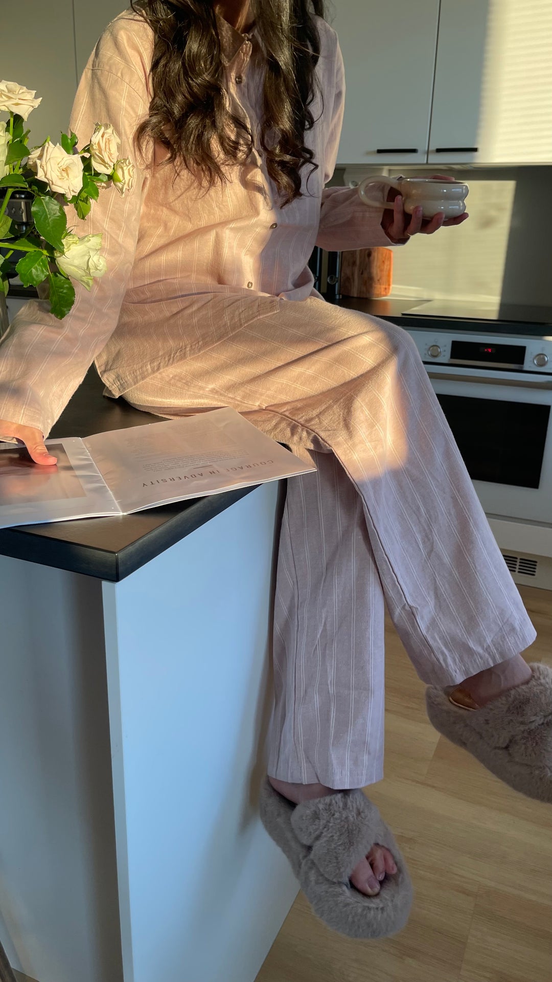 Nainen roosassa Høie Pilen -pyjamassa istuu keittiötasolla kirja kädessään
