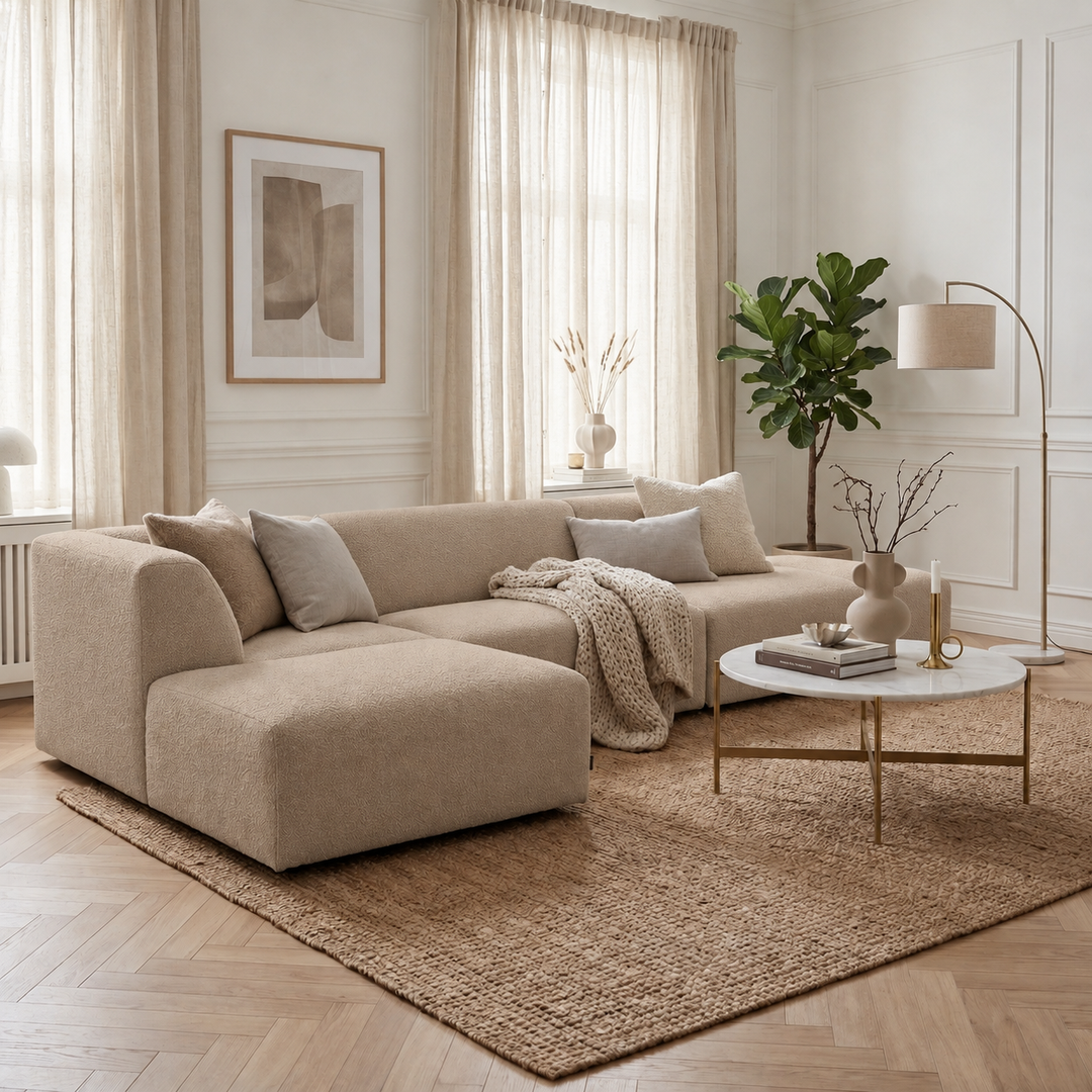 Beige Heta-divansoffa med ljusa kuddar och en stickad pläd framför ett runt vitt soffbord.