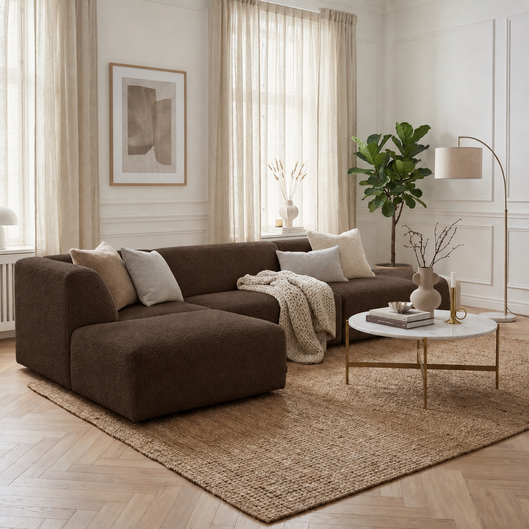 Mörkbrun Heta-divansoffa med ljusa kuddar och en stickad pläd i ett ljust rum med höga fönster.