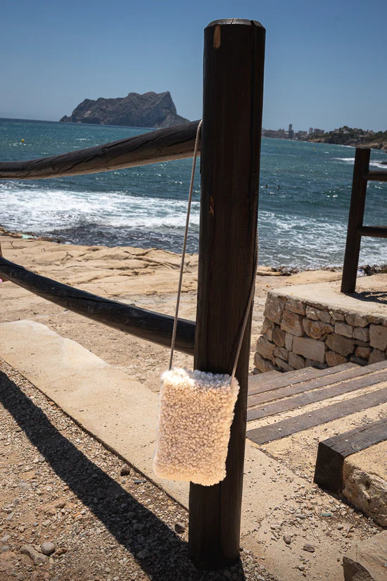 White KYWIE phone pouch hanging by its long strap from a wooden post by the sea