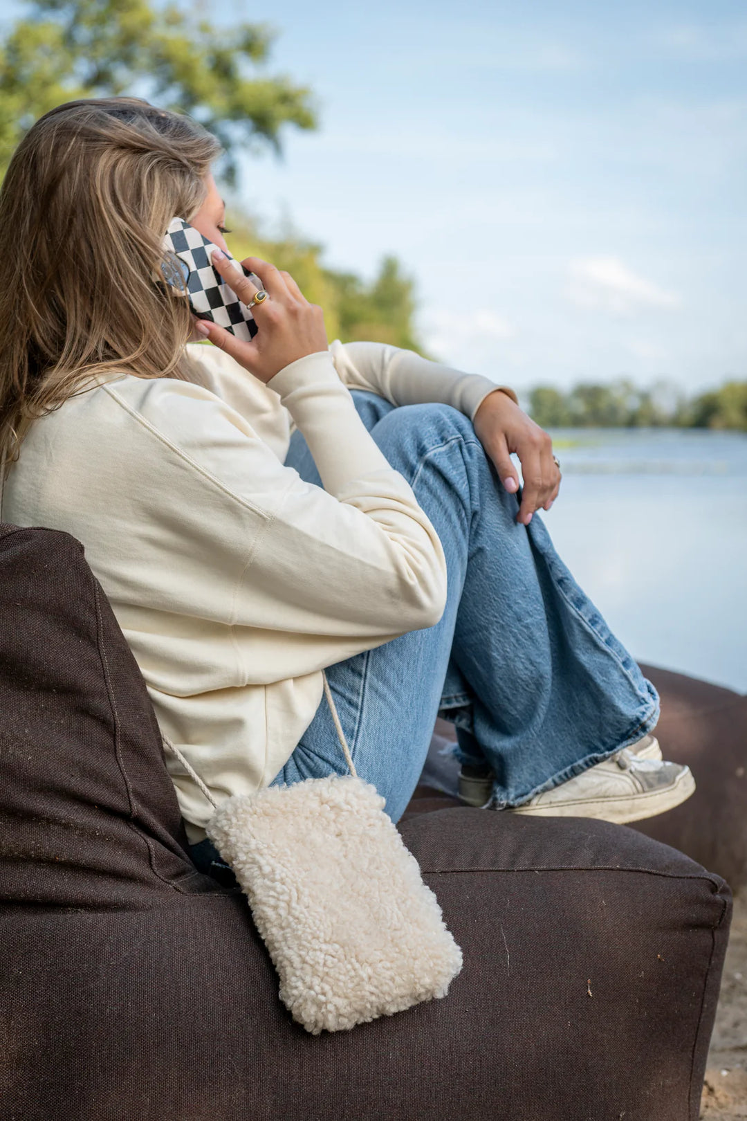 Person talking on a phone beside the water with a white KYWIE phone pouch worn across the body