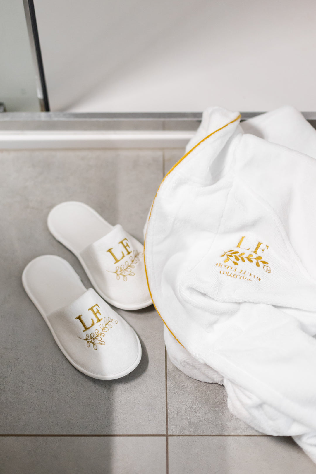 Valkoiset Loffen Hotel Luxus -kylpytossut ja kultakirjailtu kylpytakki harmaalla laattalattialla ylhäältä