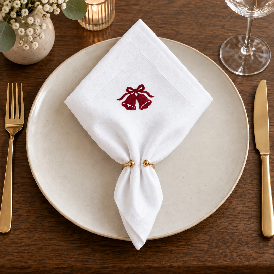 White napkin in a pointed ring fold on a plate, with red embroidered Christmas bells centred on the front.
